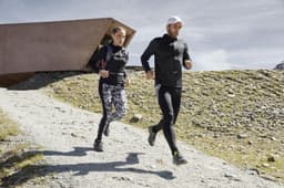 A man and woman jog outdoors wearing athletic outfits, including a black zip hoodie. The scenery is rugged and mountainous.