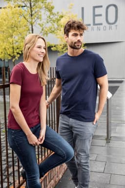 A maroon women's t-shirt and a navy men's t-shirt are worn by a model couple outdoors. Both shirts feature a logo.