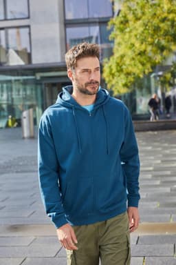 A man wearing a blue zip hoodie stands outdoors, surrounded by modern architecture and greenery.