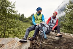 A green and blue knitted fleece vest is worn by a man, while a red vest adorns a woman. Both are outdoors in nature.
