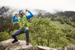 A man wearing a green and blue knitted fleece vest poses on a rock outdoors, surrounded by greenery and mountains.