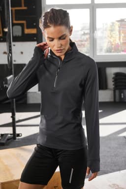 A black half-zip ladies sports shirt with long sleeves, featuring a logo, displayed in a gym setting.