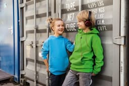 A blue hoodie and a green hoodie worn by two children, standing next to a shipping container. The children are smiling and one is wearing headphones.