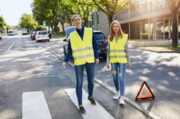 Two adults wear bright yellow safety vests while walking on a street, with a warning triangle nearby.