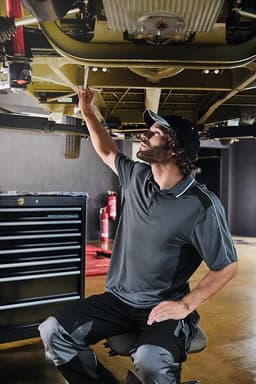 A man in a grey polo shirt works under a vehicle, using a wrench in a workshop setting with tools in the background.