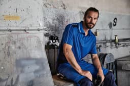 A blue polo shirt worn by a man sitting in a workshop, with tools and equipment in the background.