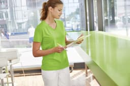 A green ladies workwear t-shirt displayed on a woman standing indoors, holding a clipboard. The setting features natural lighting.