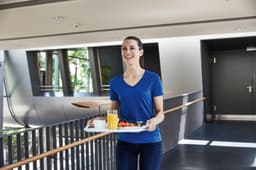 A woman in a blue V-neck t-shirt carries a tray with food and beverages in a modern interior space.