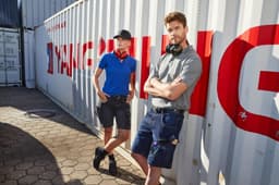 Two individuals model Workwear Stretch Bermuda Jeans in blue and grey, set against a shipping container backdrop.