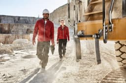 Two individuals wearing red polar fleece jackets walking in a quarry, with heavy machinery in the background.