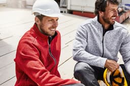 Men wearing polar fleece jackets in red and grey, sitting outdoors on a construction site. Both jackets have logos.