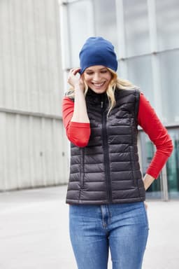A woman wears a black puffer vest over a red shirt and a blue beanie, smiling outdoors.