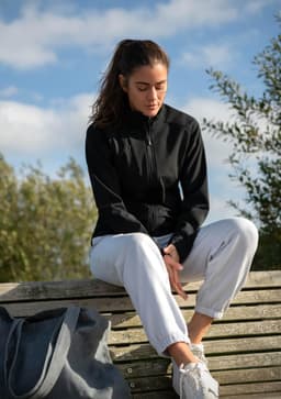 A woman sits on a bench wearing a black soft shell jacket and light trousers, with a bag beside her.