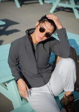 A woman in a grey soft shell jacket sits casually on a light blue table, wearing sunglasses and relaxed attire.