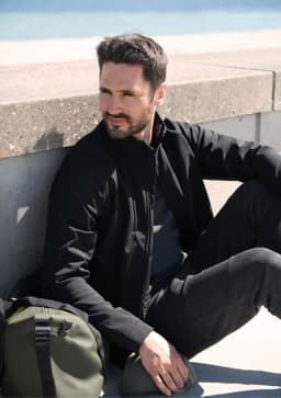 A black soft shell jacket worn by a man sitting on a ledge, with a green bag beside him.