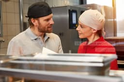 Two individuals in a kitchen setting wearing headwear, engaged in conversation, with food preparation equipment visible in the foreground.