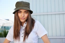 A woman wearing an olive green fisherman hat, with long hair, set against a modern building backdrop.