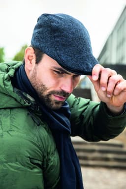 A man in a green jacket and navy scarf tips his blue cap, set against an urban backdrop.