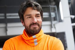 A man in an orange high-visibility shirt with reflective stripes, standing in an outdoor environment.