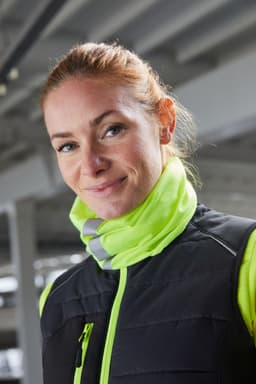 A woman wearing a black vest with a bright yellow collar. She has red hair and is smiling. The background is industrial.