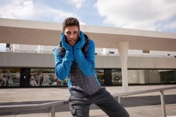 A man in a blue fleece hoodie with a grey vest stands outdoors, with a building in the background.