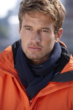 A young man wearing an orange jacket with a navy fleece scarf. The image features a blurred urban background.
