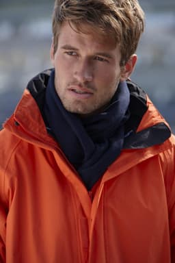 A man wearing an orange jacket and a navy fleece scarf, positioned outdoors with a blurred background.
