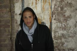 A person wearing a grey fleece scarf and a black hoodie stands against a weathered brick wall.