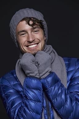 A man in a grey beanie and scarf, smiling, wearing a blue puffer jacket. The image features a logo on the scarf.