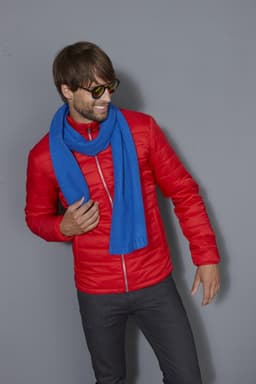 A man in a red jacket wears a blue scarf. He is smiling and posing against a grey background.