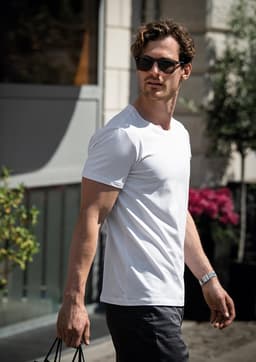 A man wearing a white T-shirt and sunglasses, walking outdoors with a bag in hand.