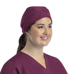 A unisex scrub cap in a rich plum colour with buttons, worn by a smiling woman in a matching top.