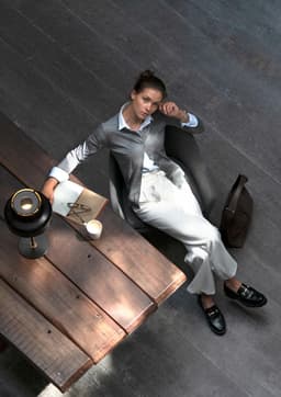 A woman in a grey cardigan sits on a wooden table, wearing light trousers and black shoes, with a lamp and notebook nearby.