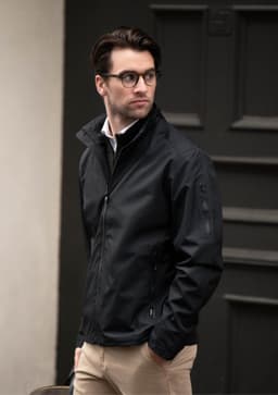 A man wearing a black jacket stands outdoors, paired with light trousers and glasses, against a dark background.