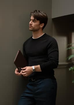 A black knitted jumper worn by a man leaning against a wall, holding a brown folder in his hand.