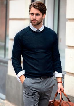 A man wearing a navy knitted jumper over a collared shirt, paired with grey trousers and holding a brown leather bag.