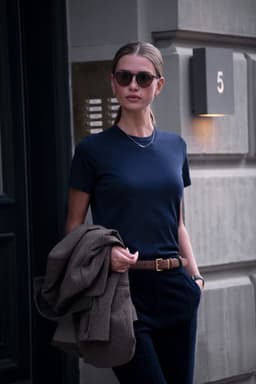 A woman wearing a navy blue t-shirt, holding a brown jacket and wearing sunglasses, stands in front of a building.