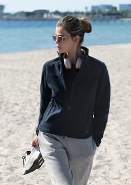 A women’s navy jacket with a high collar, paired with grey pants, worn by a model on a sandy beach.