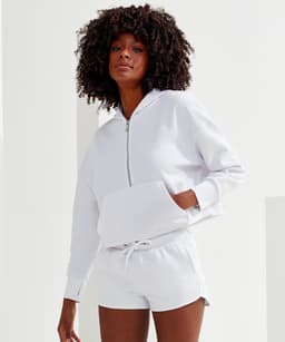 A white 1/2 zip hoodie featuring a front pocket, styled with matching shorts. The model has curly hair.
