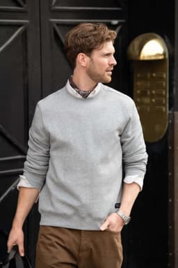 A grey unisex sweatshirt worn by a man, paired with a collared shirt and brown trousers, featuring a logo.