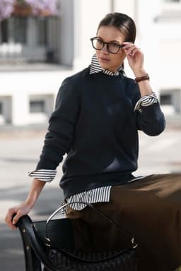 A dark grey unisex sweatshirt worn over a striped shirt, paired with a long skirt and accessorised with glasses.