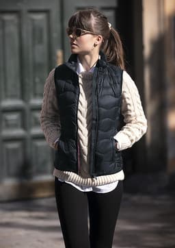 A woman wearing a black puffer vest over a cream knitted sweater, standing outdoors with hands in pockets.