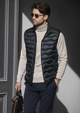 A man wearing a black puffer vest over a light grey turtleneck, holding a brown leather bag.