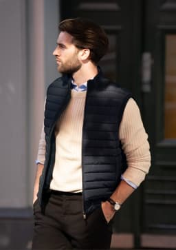 A man wearing a navy puffer vest over a cream sweater, standing outside with a stylish backdrop.