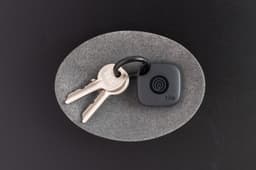A key ring with a grey Tile Mate tracker attached, resting on a circular grey surface.