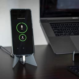 A transparent phone stand displaying a charging smartphone beside a laptop on a desk.