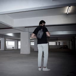 A man in a black shirt and light trousers is carrying a grey anti-theft sling bag in a parking garage.