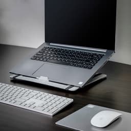 A grey laptop stand with a laptop, white keyboard, and mouse on a dark wooden desk.