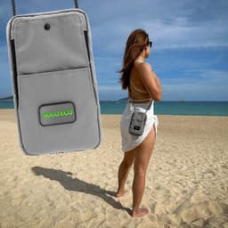 A grey eco-friendly bag displayed on a beach, featuring a logo, with a person wearing it while facing the ocean.