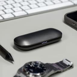 A black charging case for true wireless earbuds rests on a desk next to a watch, a smartphone, and a pen.